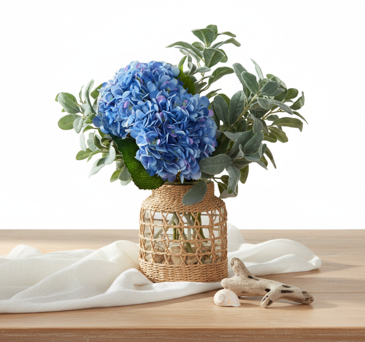 An artificial floral arrangement featuring a large blue hydrangea bloom, green and silver eucalyptus leaves, and a woven natural fiber basket vase.