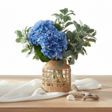 An artificial floral arrangement featuring a large blue hydrangea bloom, green and silver eucalyptus leaves, and a woven natural fiber basket vase.