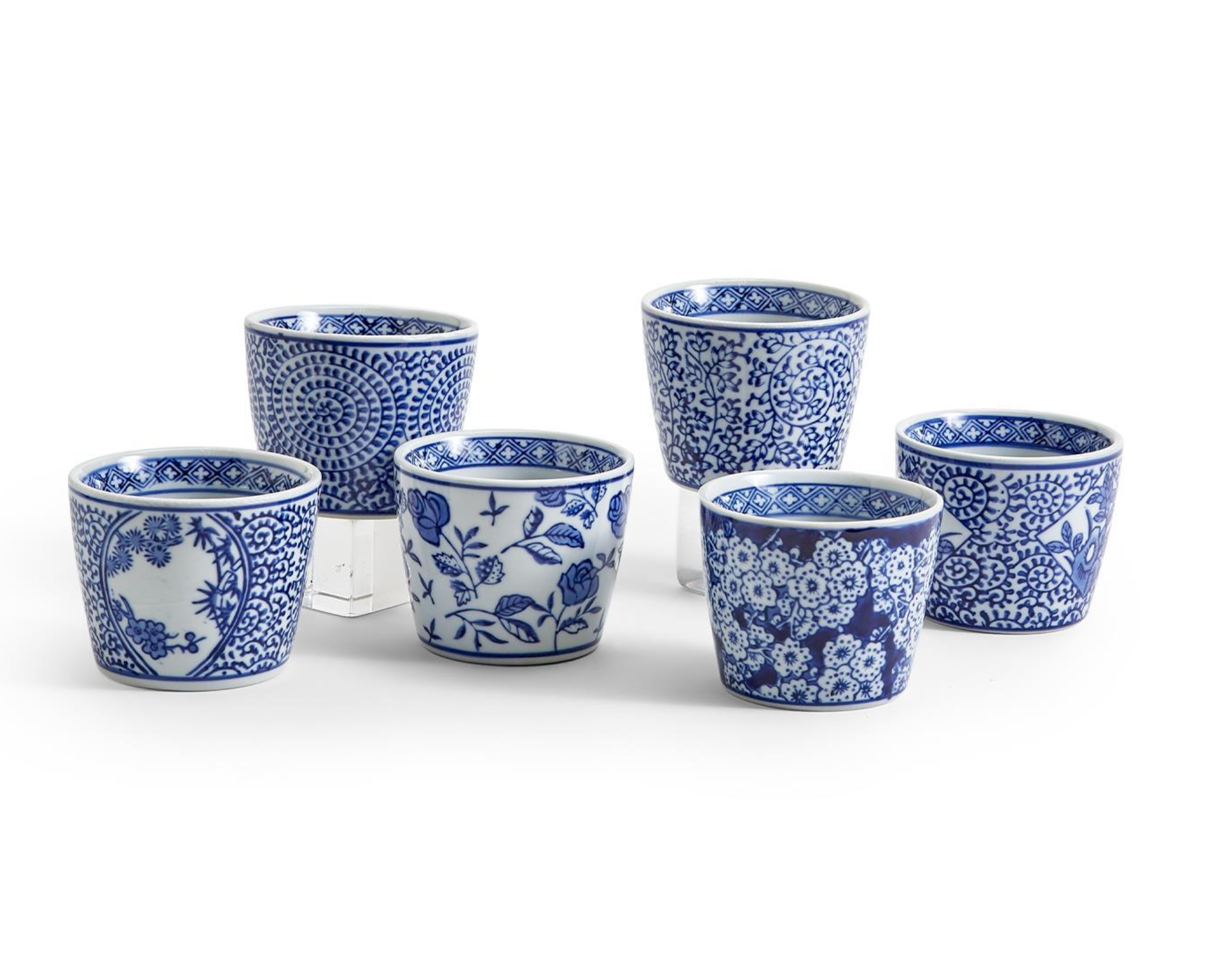 Six blue and white patterned ceramic cups arranged on a white background, with three taller cups behind three shorter cups. The cups feature various floral and decorative blue motifs.