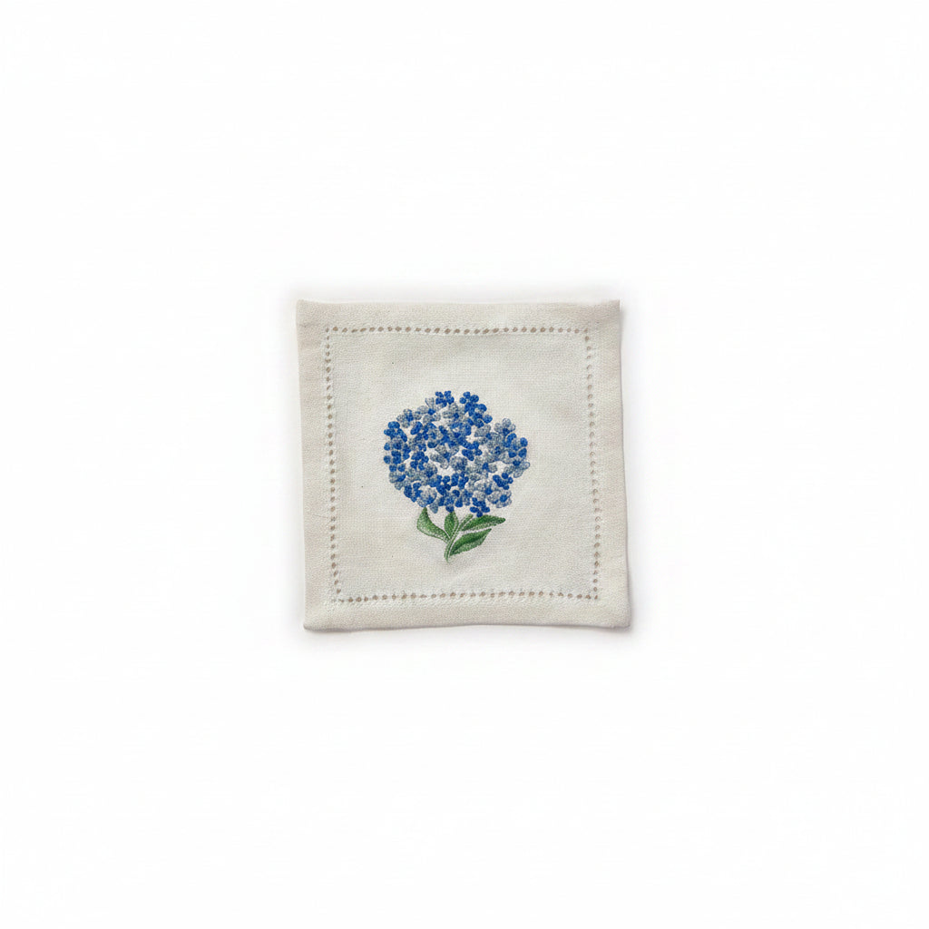 A white cotton cocktail napkin with hand embroidered blue hydrangea flowers and green leaves.