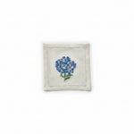 A white cotton cocktail napkin with hand embroidered blue hydrangea flowers and green leaves.