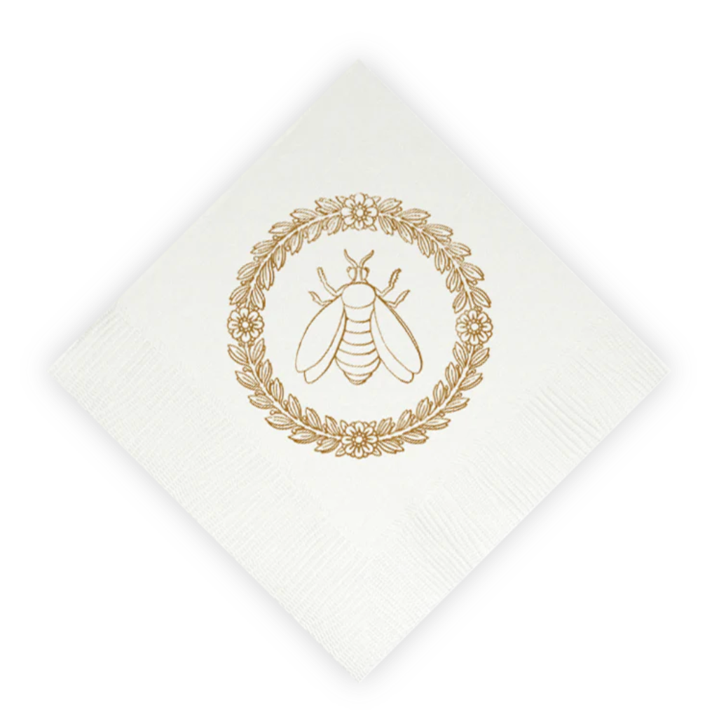 A white paper napkin, rotated at a 45-degree angle, featuring a gold foil stamp of a bumblebee inside a circular leafy wreath.