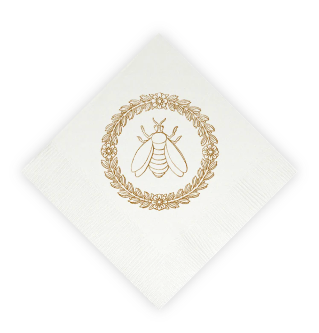A white paper napkin, rotated at a 45-degree angle, featuring a gold foil stamp of a bumblebee inside a circular leafy wreath.