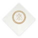 A white paper napkin, rotated at a 45-degree angle, featuring a gold foil stamp of a bumblebee inside a circular leafy wreath.