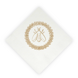 A white paper napkin, rotated at a 45-degree angle, featuring a gold foil stamp of a bumblebee inside a circular leafy wreath.