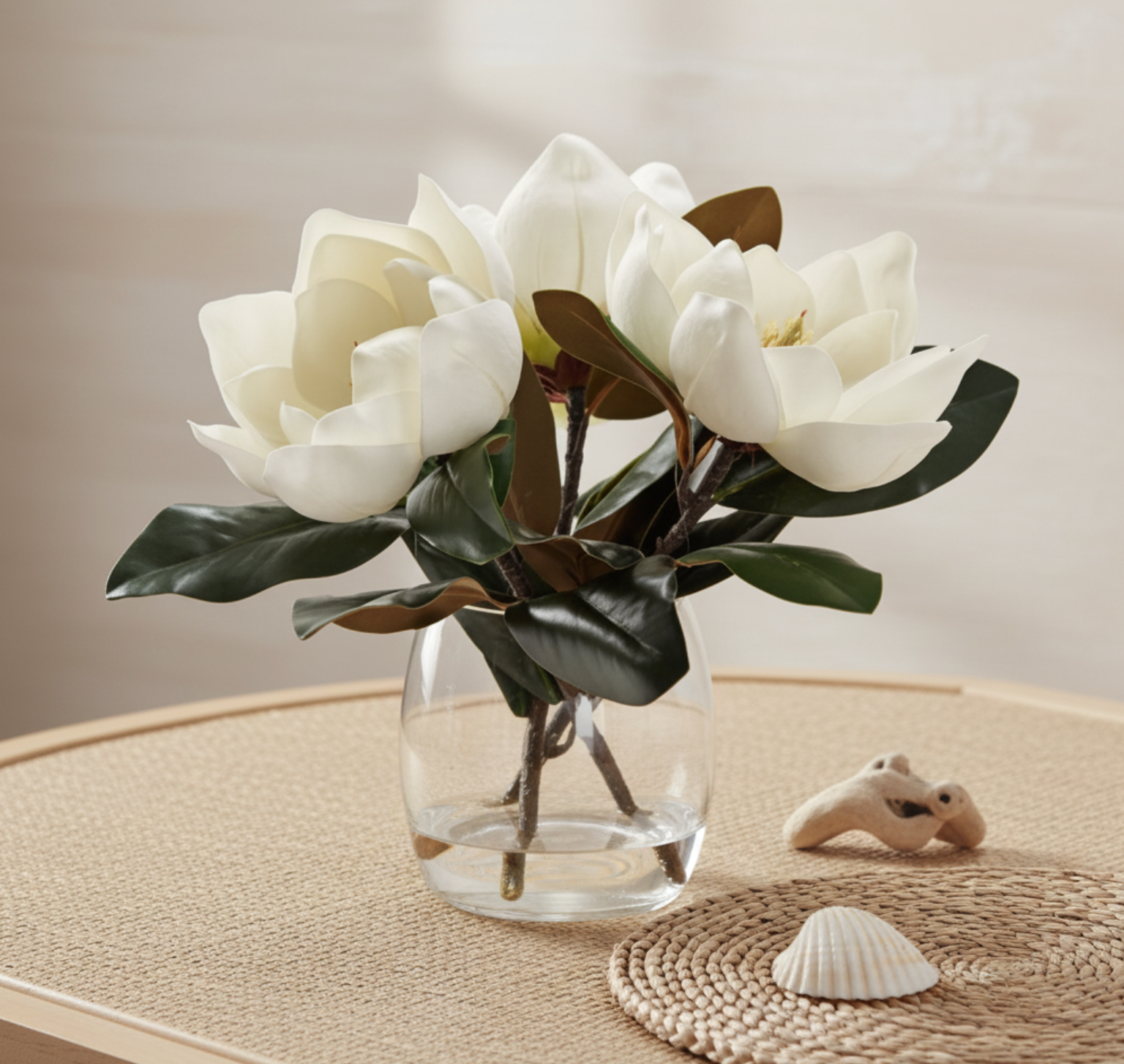 White magnolia artificial flowers with dark green leaves arranged in a clear glass vase.
