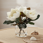 White magnolia artificial flowers with dark green leaves arranged in a clear glass vase.