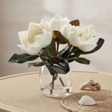 White magnolia artificial flowers with dark green leaves arranged in a clear glass vase.