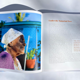 An open printed book or magazine spread photographed against a light gray background. The left page shows a color photograph of an elderly woman in profile wearing a white headscarf and purple garment, with blue sky and plants in the background.