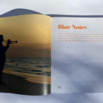 An open book or magazine spread photographed outdoors against a cloudy sky. The left page shows a large photo of a silhouetted person playing a trumpet on a beach at sunset, and the right page has the headline 'Blue Notes' with body text and a large block of smaller text.