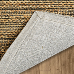 A close-up of a woven area rug corner showing its light gray underside, placed over a natural tan and brown woven rug.