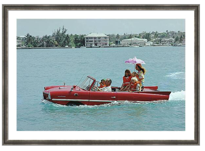 Framed wall art print showing a red convertible speedboat with several people on the water, with a distant shoreline of houses and trees under a pale sky. The print is presented in a white mat and a dark wood frame.