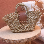 A woven straw tote bag with a brown leather handle and a decorative panel of brass studs.