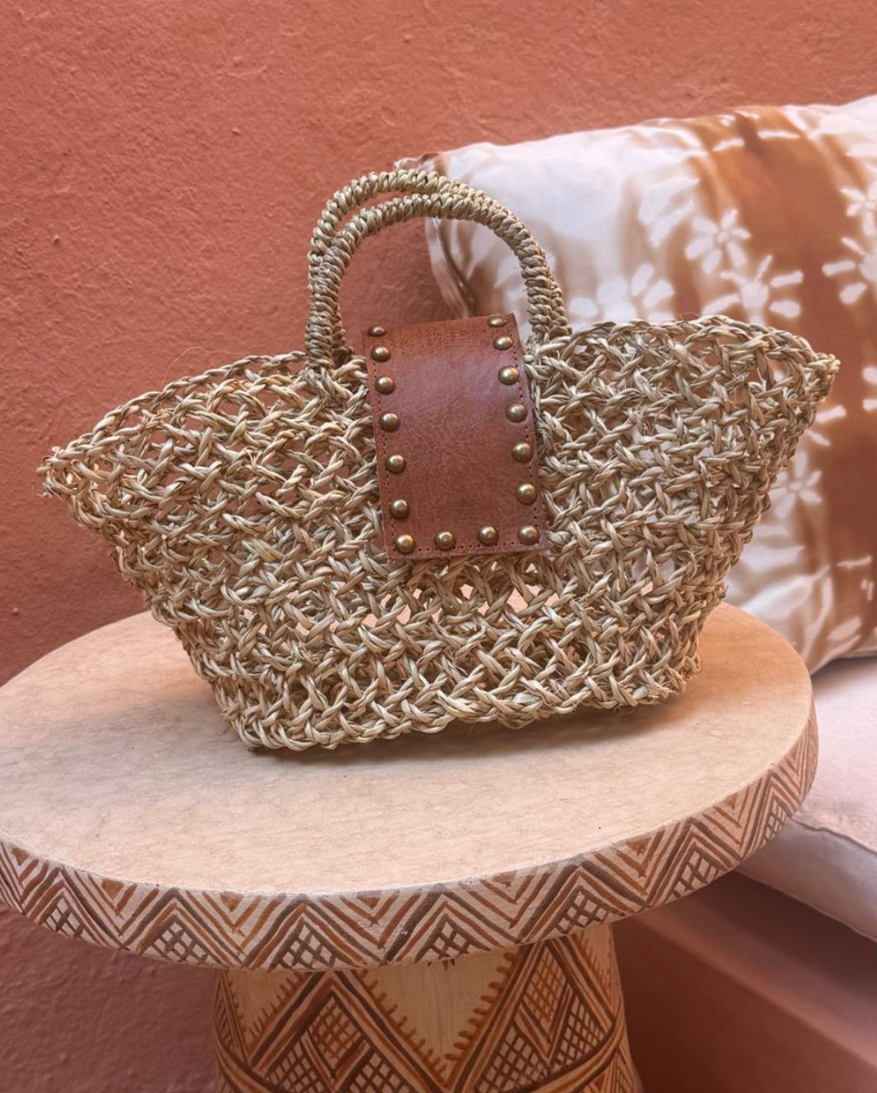 A woven straw tote bag with a brown leather handle and a decorative panel of brass studs.