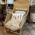 A natural wicker armchair styled with a cream throw blanket, patterned pillows, and a fringed accent, set on a textured rug inside a home decor store.