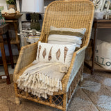A natural wicker armchair styled with a cream throw blanket, patterned pillows, and a fringed accent, set on a textured rug inside a home decor store.