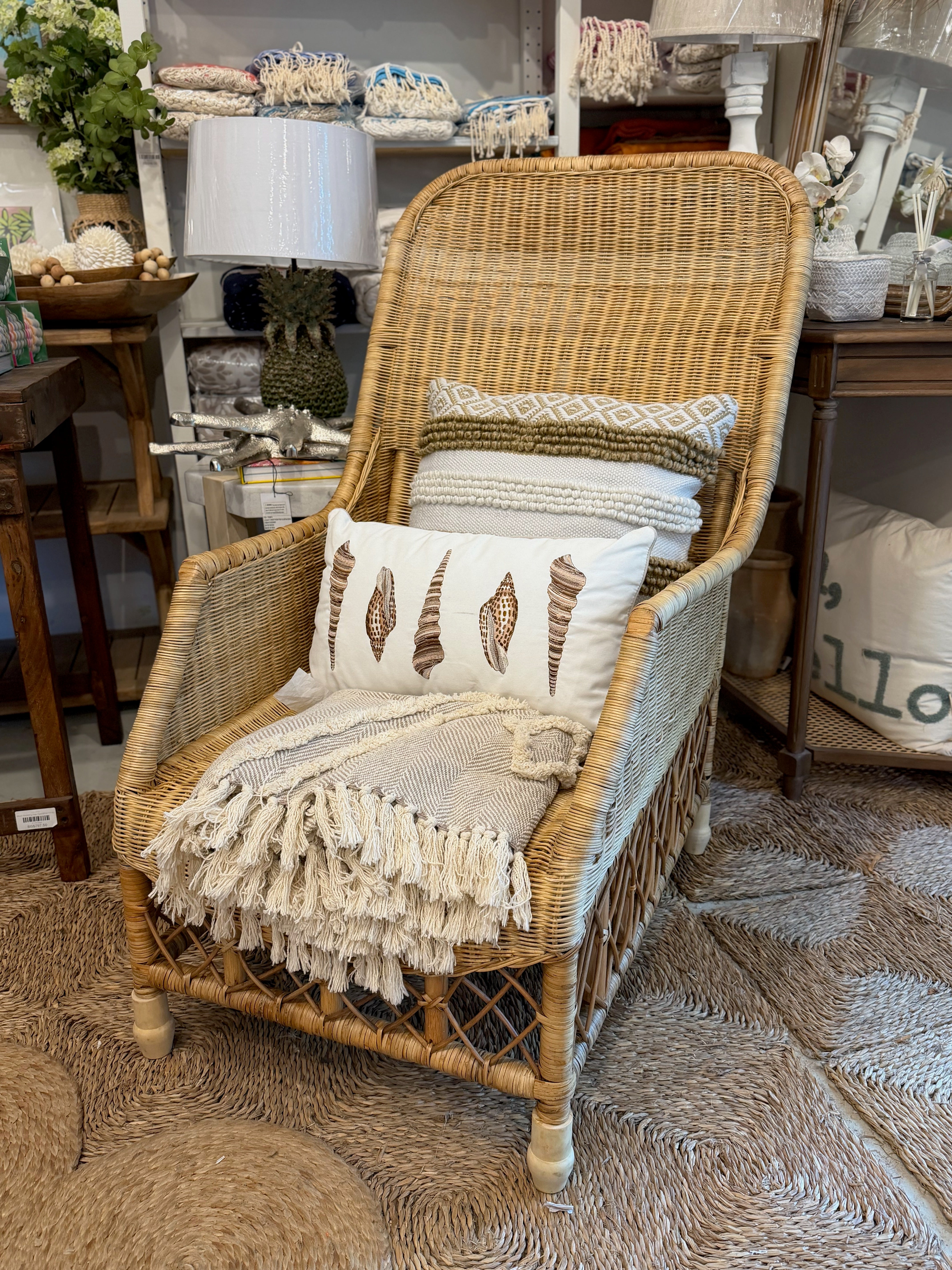 A natural wicker armchair styled with a cream throw blanket, patterned pillows, and a fringed accent, set on a textured rug inside a home decor store.