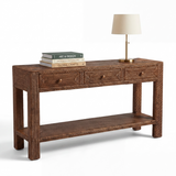 A dark brown woven rattan console table with three drawers and an open lower shelf, styled with a stack of books and a table lamp on a white background.