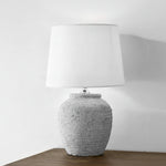 A white ceramic lamp with a rough texture, featuring a cylindrical shape and a white lampshade, placed on a flat surface with a neutral background.