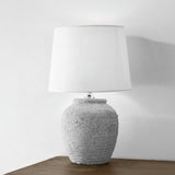 A white ceramic lamp with a rough texture, featuring a cylindrical shape and a white lampshade, placed on a flat surface with a neutral background.