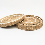 Two round woven seagrass or straw placemats with a natural tan color and white braided accents.