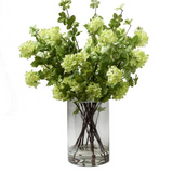 An artificial flower bouquet with light green hydrangea-like blooms and green leaves, arranged in a clear cylindrical glass vase.