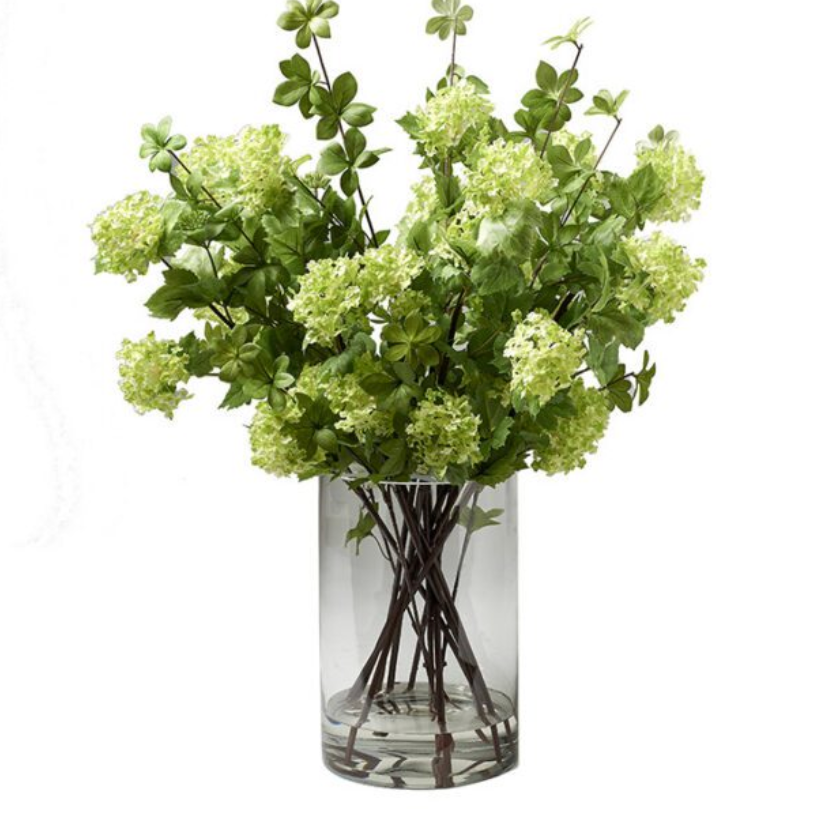 An artificial flower bouquet with light green hydrangea-like blooms and green leaves, arranged in a clear cylindrical glass vase.