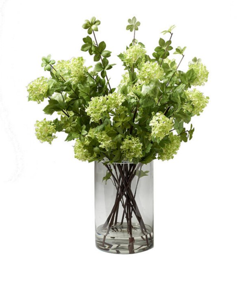 An artificial flower bouquet with light green hydrangea-like blooms and green leaves, arranged in a clear cylindrical glass vase.