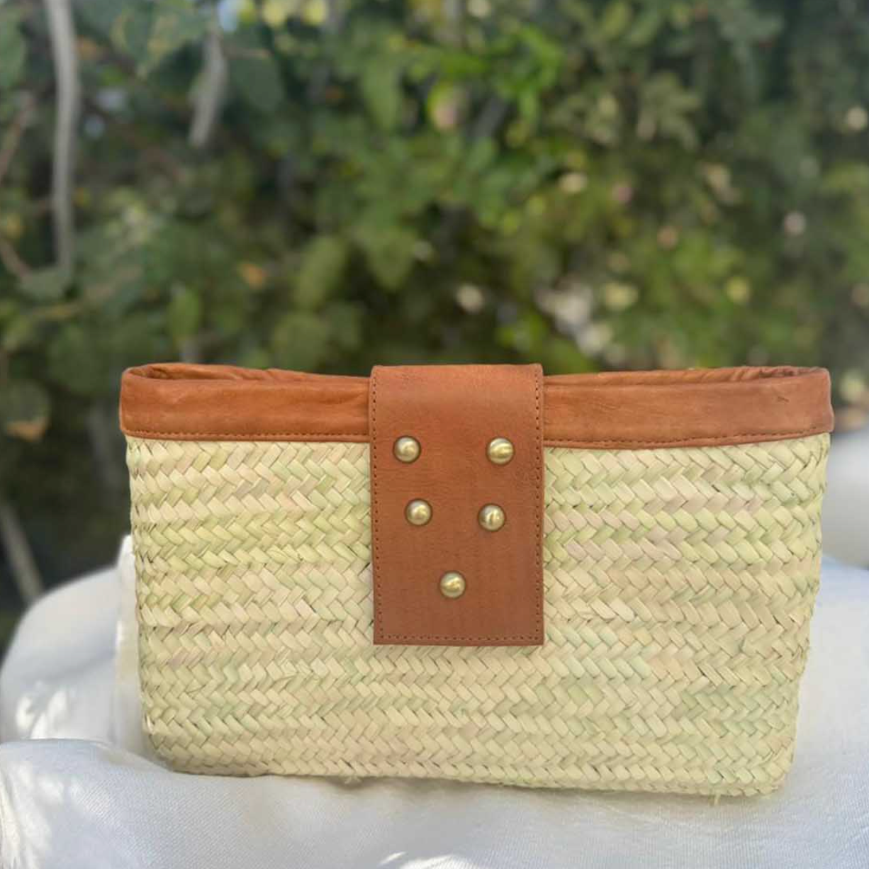 A woven straw clutch bag with a tan leather strap and gold-tone studs.