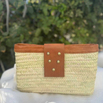 A woven straw clutch bag with a tan leather strap and gold-tone studs.