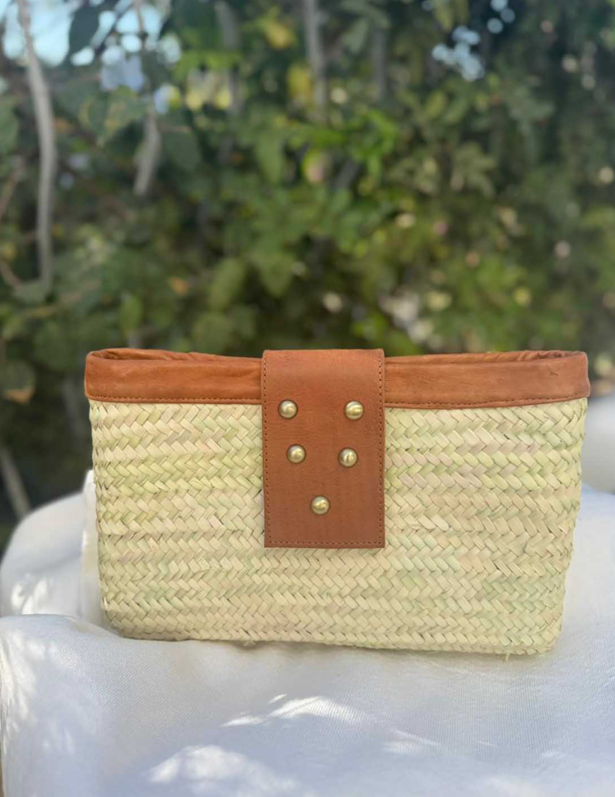 A woven straw clutch bag with a tan leather strap and gold-tone studs.