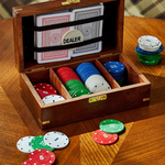 An open wooden poker box on a wooden table, filled with playing cards and stacks of colored chips. A round dealer badge is visible inside the lid, and additional chips are scattered around the box.