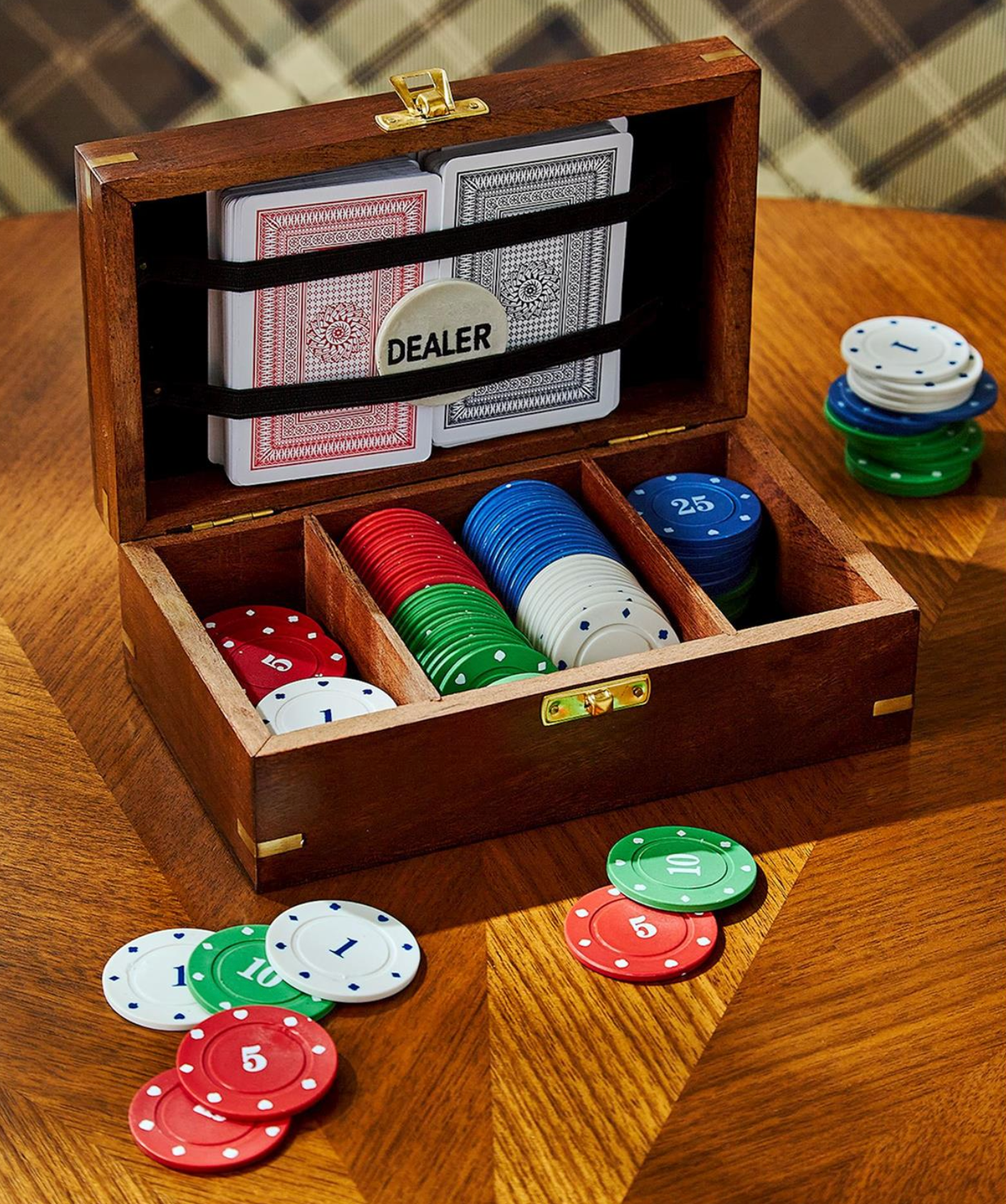 An open wooden poker box on a wooden table, filled with playing cards and stacks of colored chips. A round dealer badge is visible inside the lid, and additional chips are scattered around the box.
