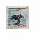 A framed painting of a sea turtle swimming in blue water with sandy shore details, set in a distressed white wood frame 