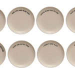 Eight round ceramic coasters in a warm off-white color, arranged in two rows of four on a plain white background. Each coaster features a different humorous phrase printed in black sans-serif font.
